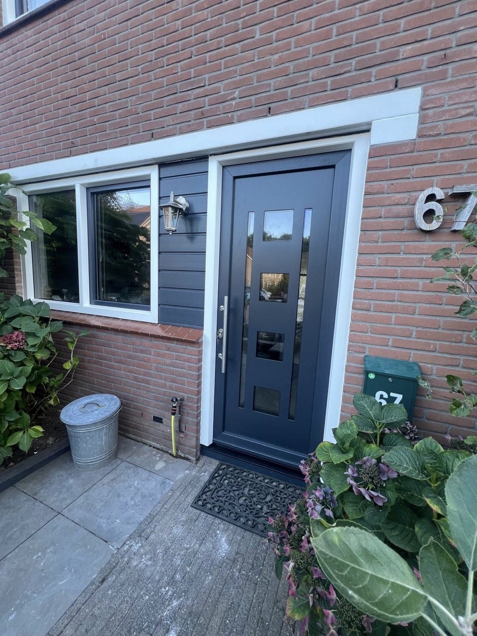 De antraciete voordeur met glasvlakken en lange trekker, naast het raam en de donkere gevelbekleding. Projectfoto.